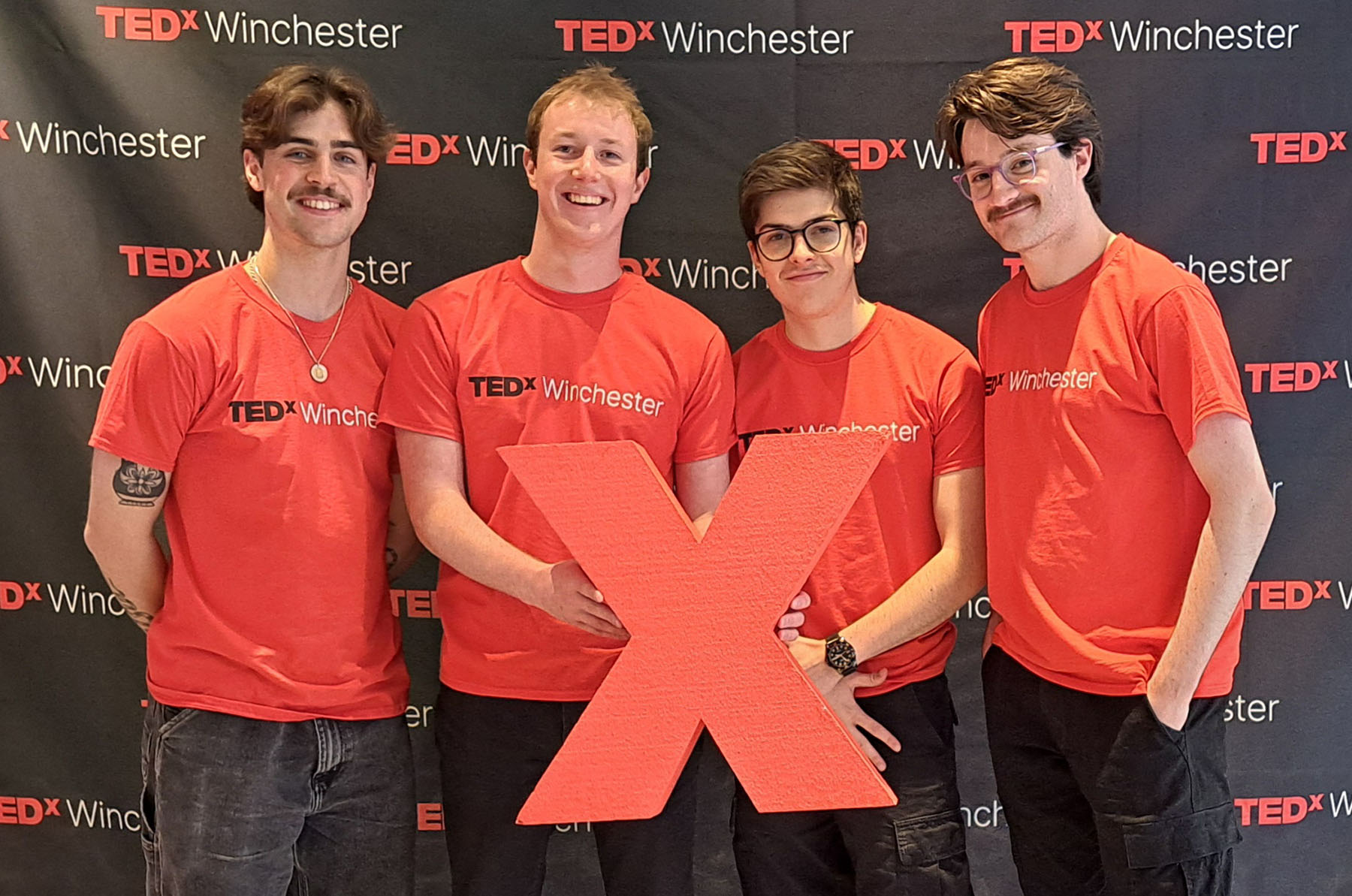 four smiling young men in red TEDx t-shirts holding a large red X